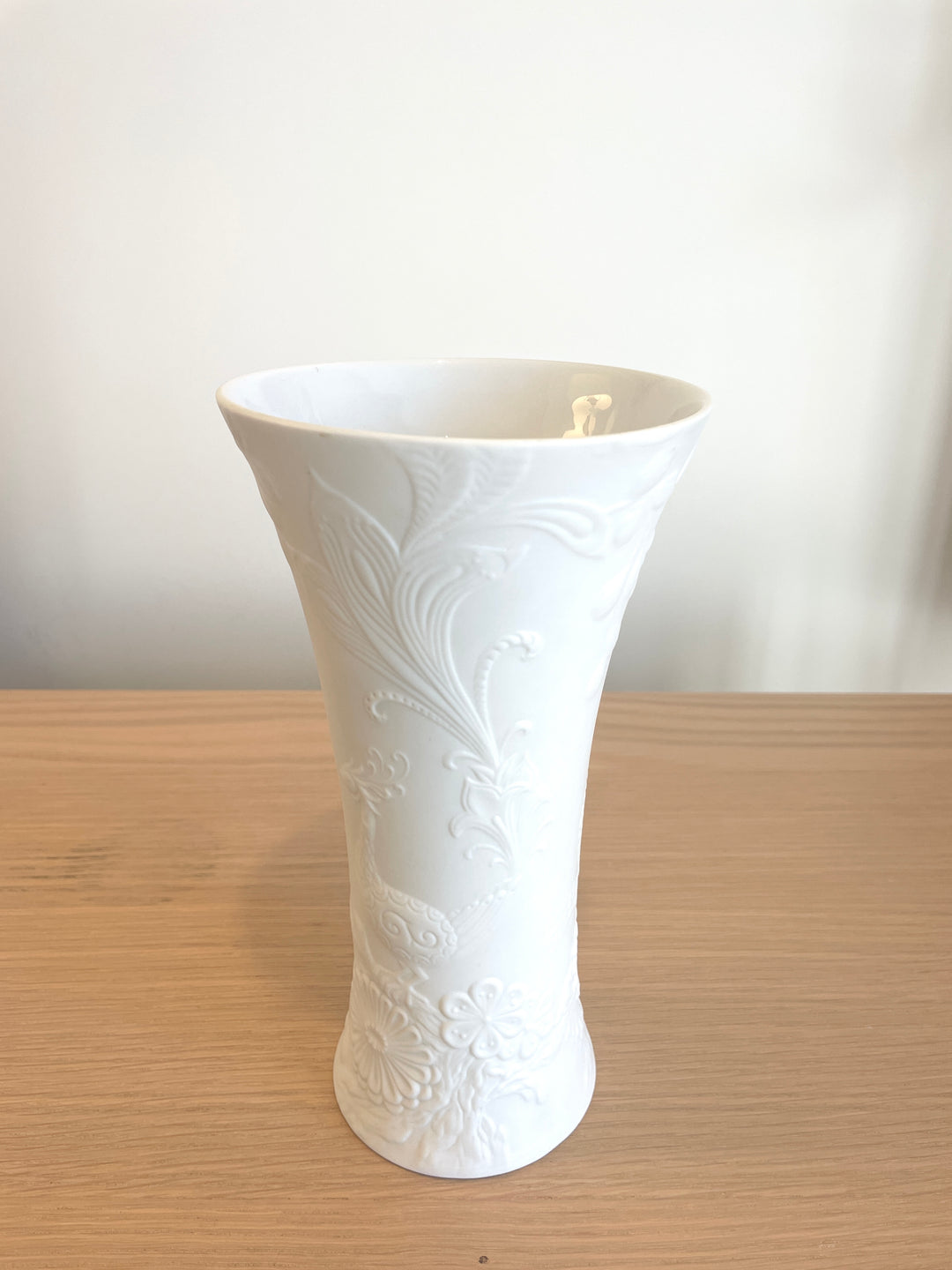White porcelain vase by Kaiser, featuring raised floral and peacock designs, standing on a wooden surface.