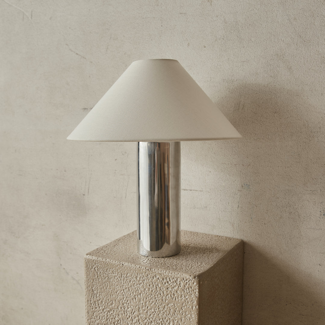 Chrome table lamp by McMullin, with a simple white lampshade, on a textured beige pedestal.
