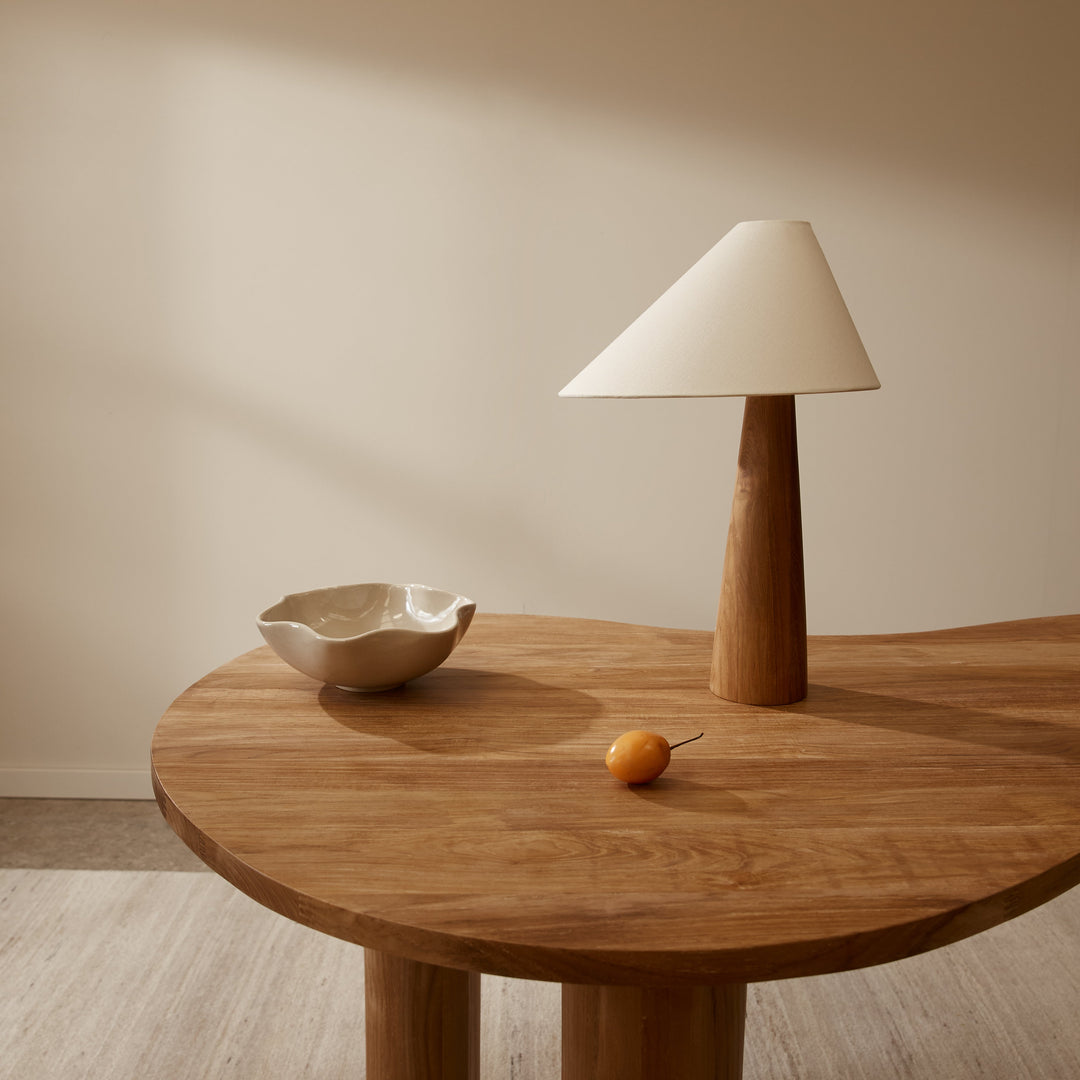 Table lamp from McMullin with a wood base and a triangular white shade on a wooden table with a bowl and orange.