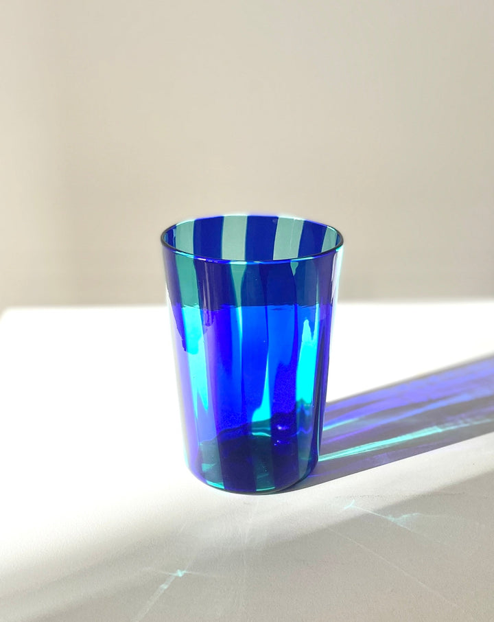 Striped blue drinking glass by Shop Dessein Parke with refracted light on a white surface.
