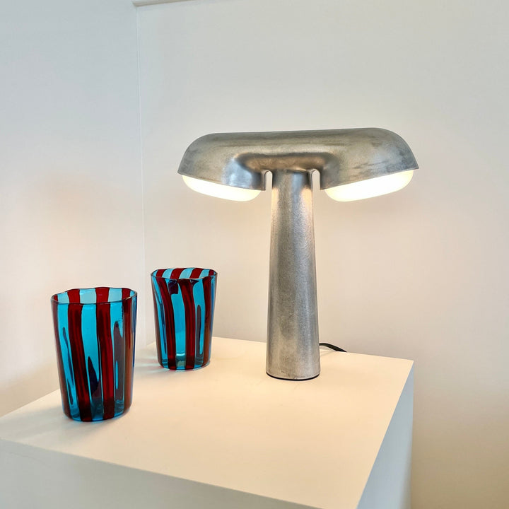 Raw metal TGV table lamp by Moustache Paris, with two lit domes, sitting next to blue and red striped glasses.