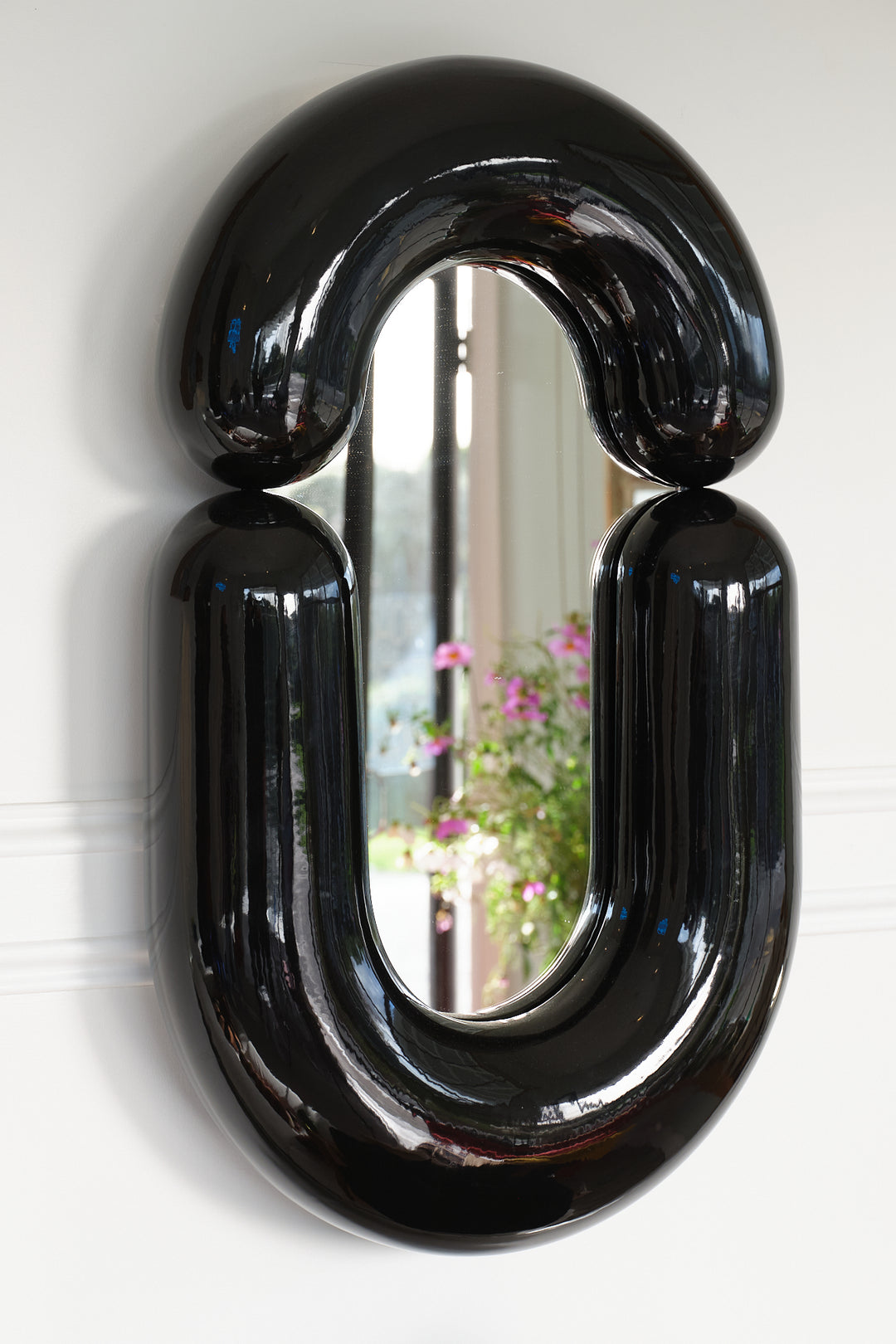 Black wall mirror by Moustache Paris with a distinctive rounded shape, reflecting a garden view.
