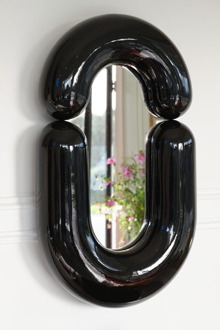 Black wall mirror by Moustache Paris with a distinctive rounded shape, reflecting a garden view.
