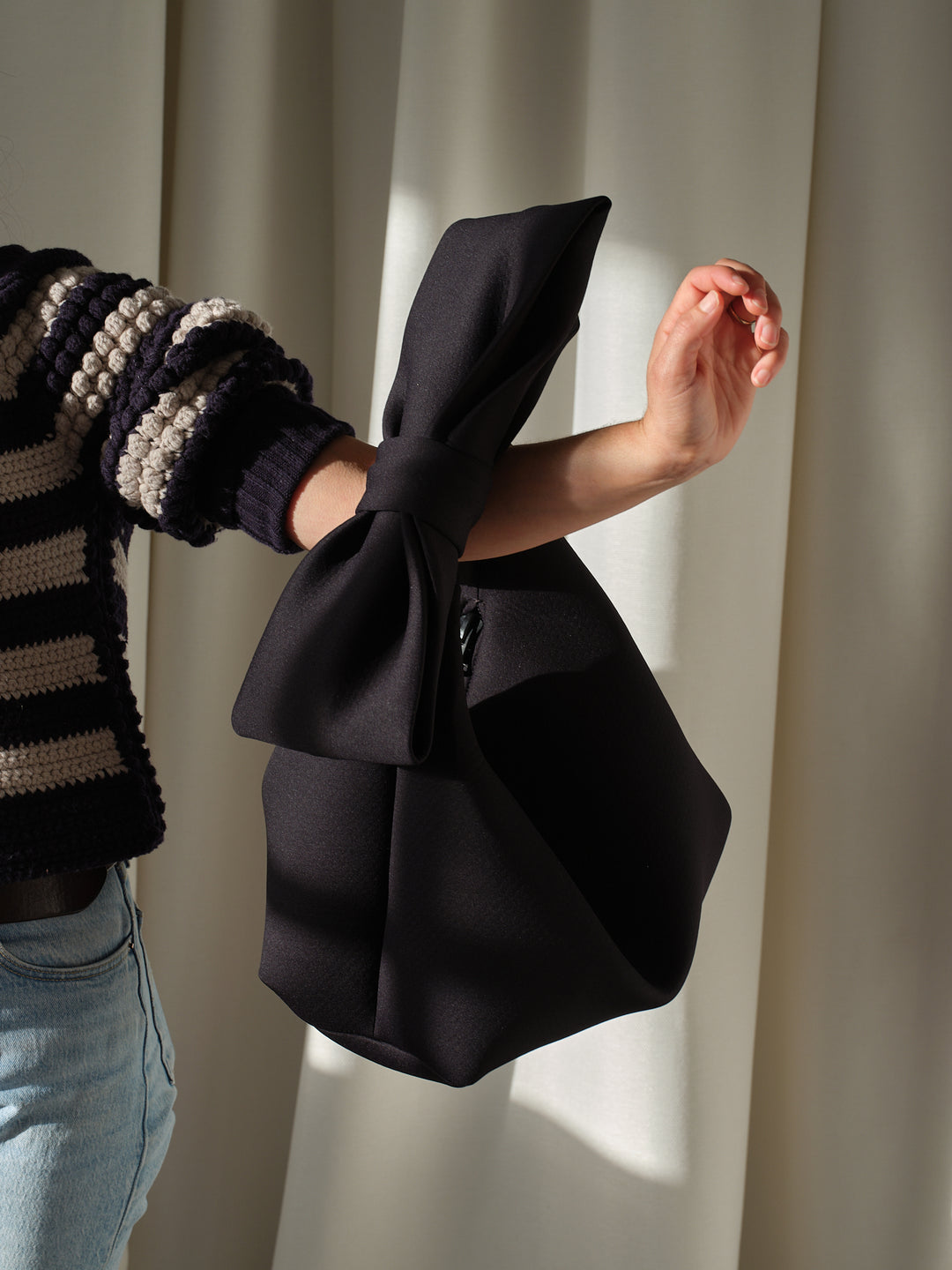 Black Jaimie Bow handbag from Kara, held on an arm with striped sweater and jeans against curtain background.