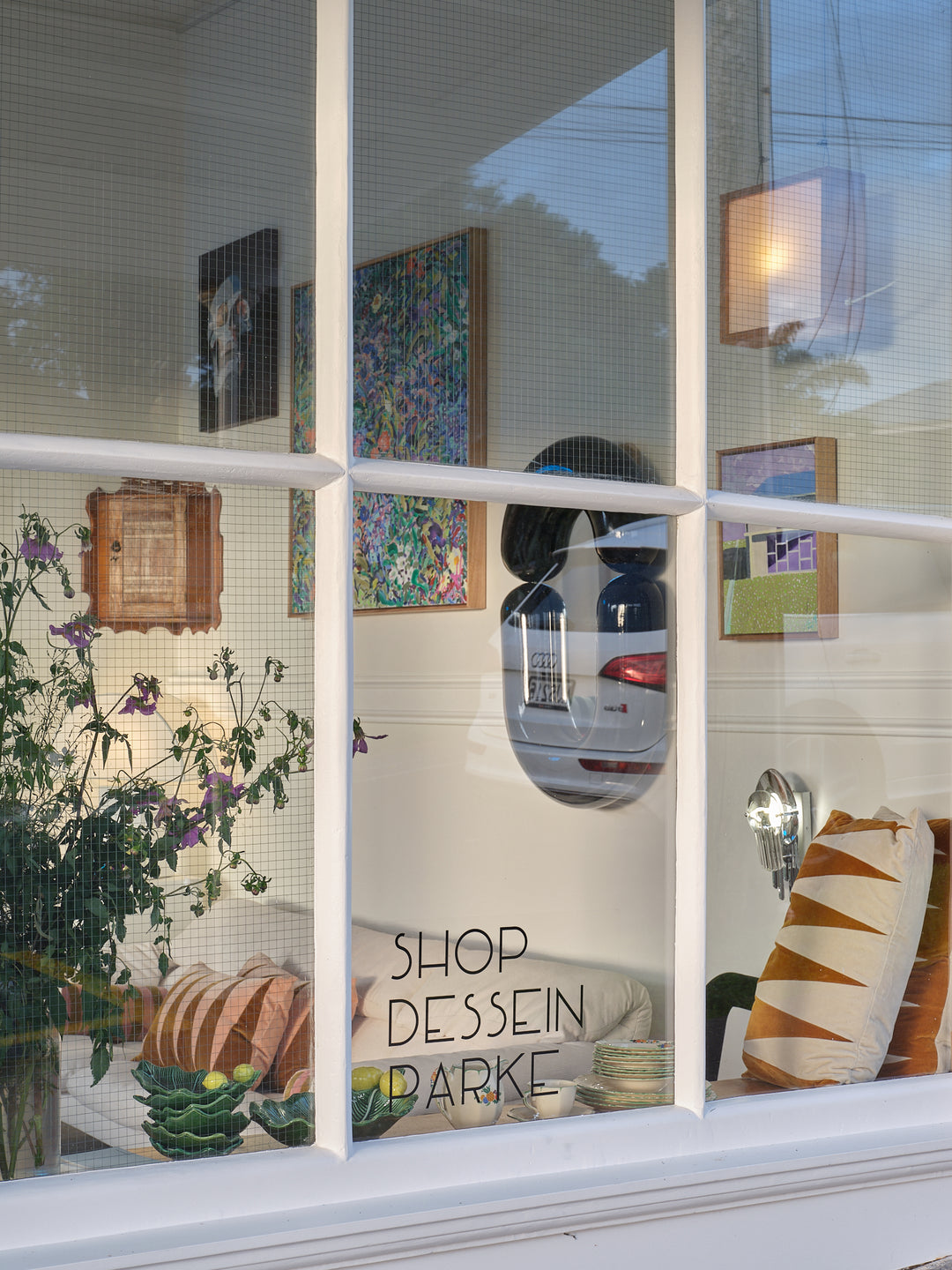 Window display with plants, pillows, and decor, reflecting a car on the glass.
