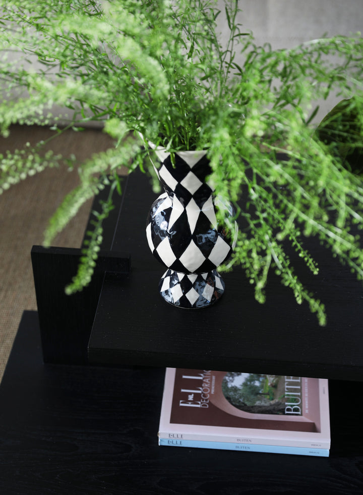 Black and white patterned rhombic vase by &Klevering holding green leafy plants, sitting on a black table with magazines underneath.
