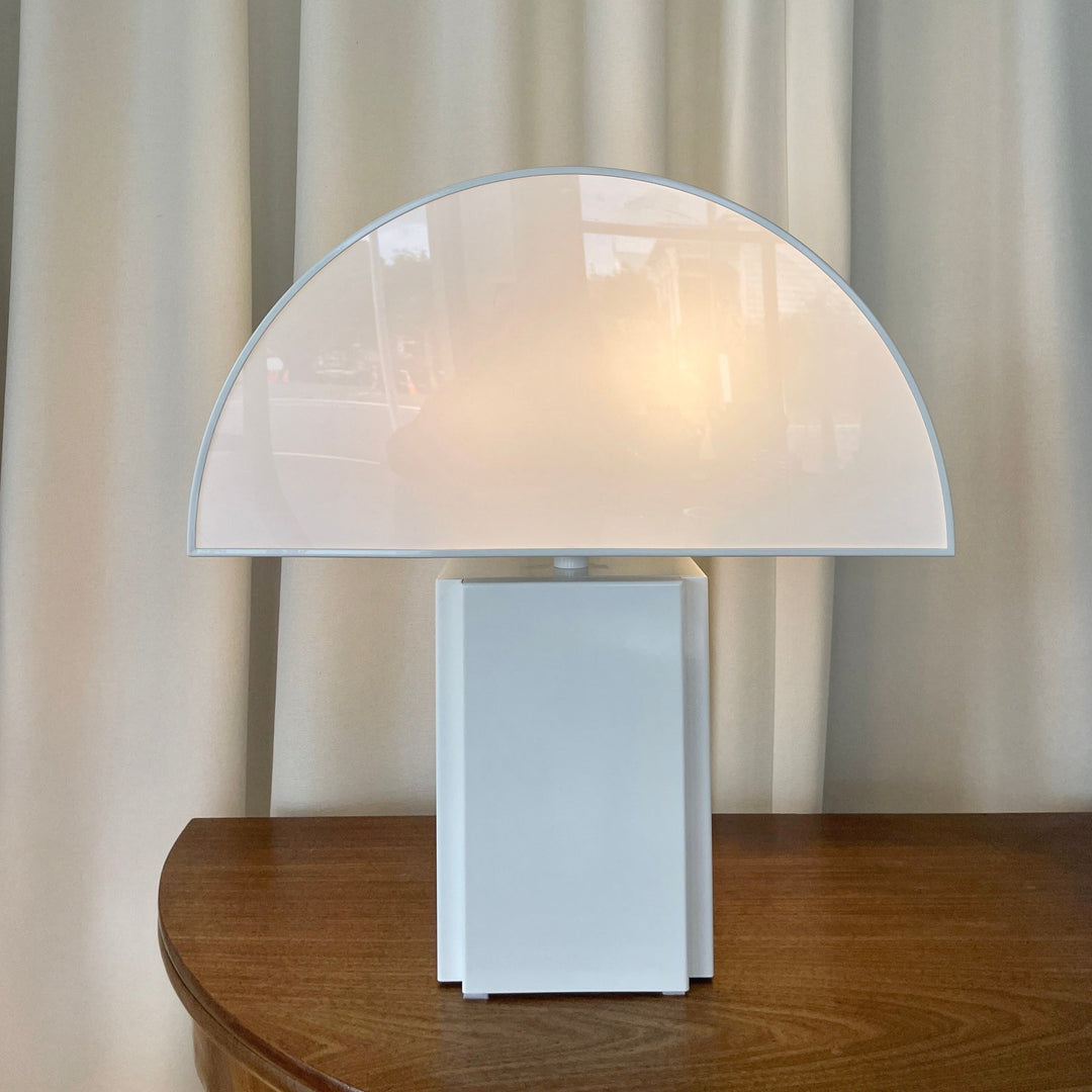 White table lamp by Harvey Guzzini, with a semi-circular shade, sitting on a wooden table.