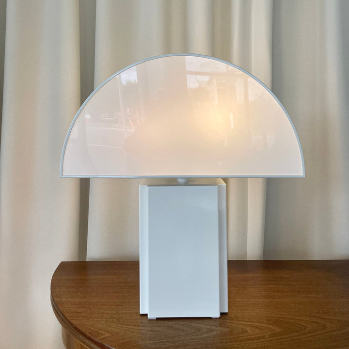 White table lamp by Harvey Guzzini, with a semi-circular shade, sitting on a wooden table.