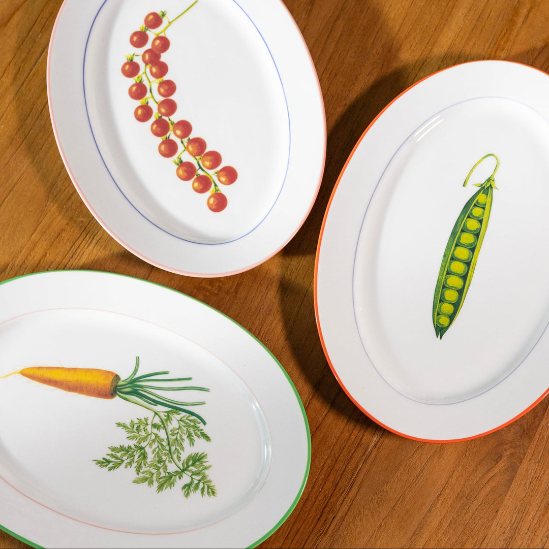 White ceramic serving plates by &Klevering with carrot, pea, and cherry tomato illustrations, arranged on a wooden surface.