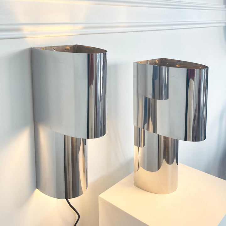 Stainless steel wall and table light from Edera Radice, with geometric shape, against a white wall.