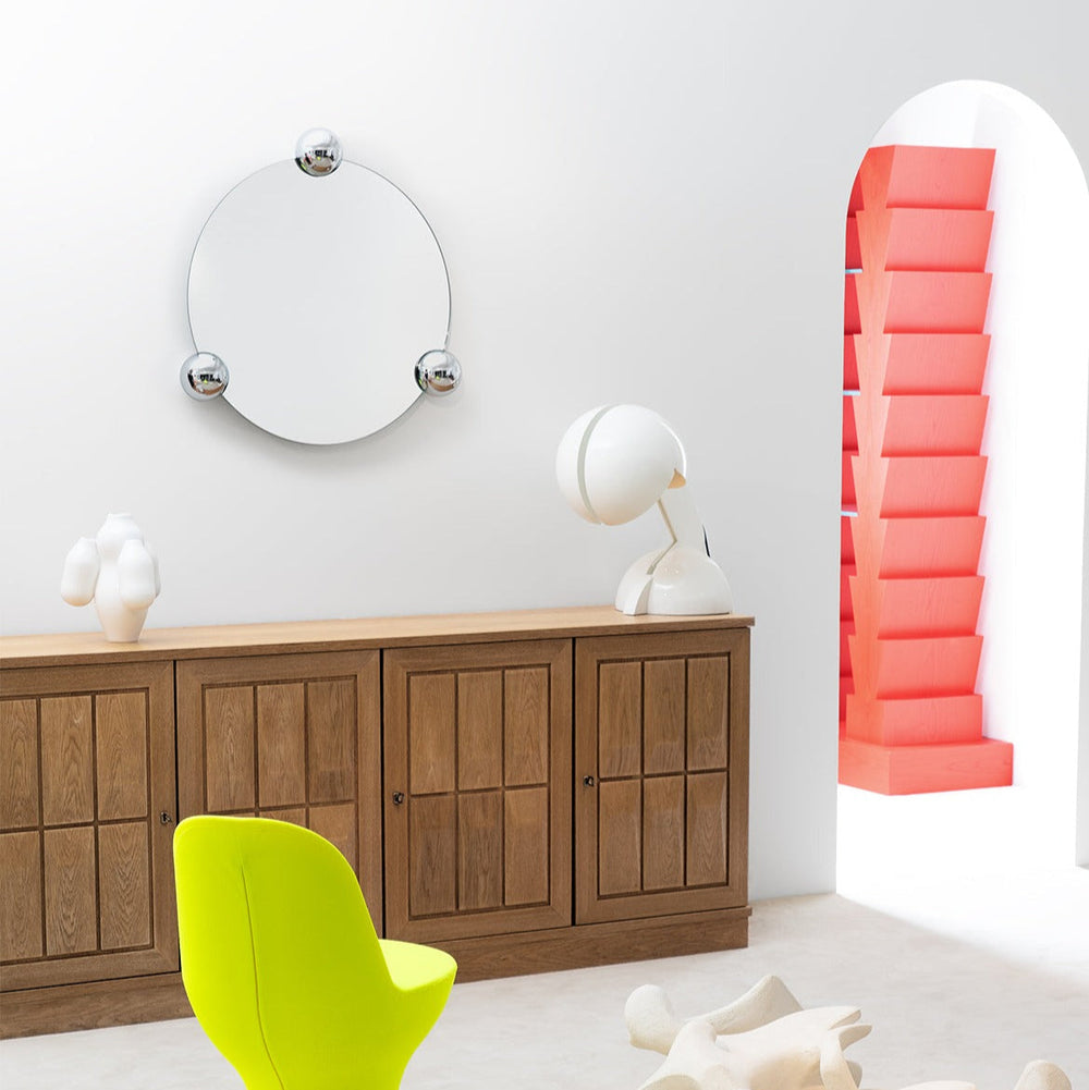Round wall mirror by Moustache Paris with three silver ball accents, hanging above a wooden credenza.
