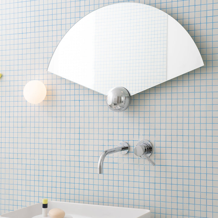 Fan-shaped Sphera wall mirror by Moustache Paris, reflecting a grid-patterned wall in a contemporary bathroom.
