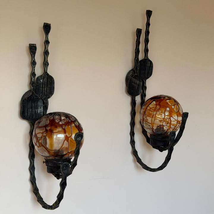 A pair of decorative wall sconces with dark twisted metal arms and oval glass shades featuring a mottled amber-brown and cream pattern, mounted on a plain beige wall.