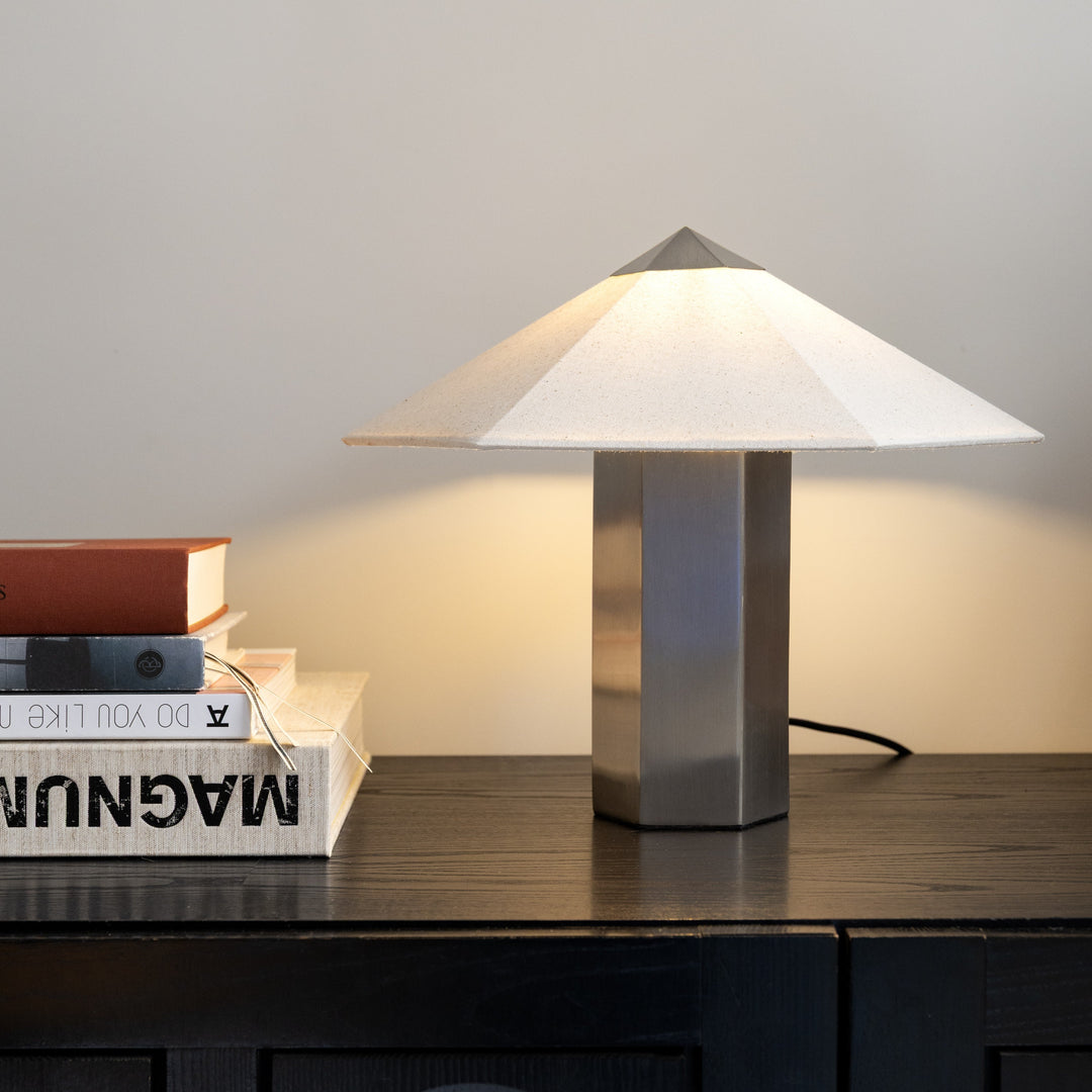 Brushed stainless steel table lamp by &Klevering with a linen lampshade on a wooden table with books.