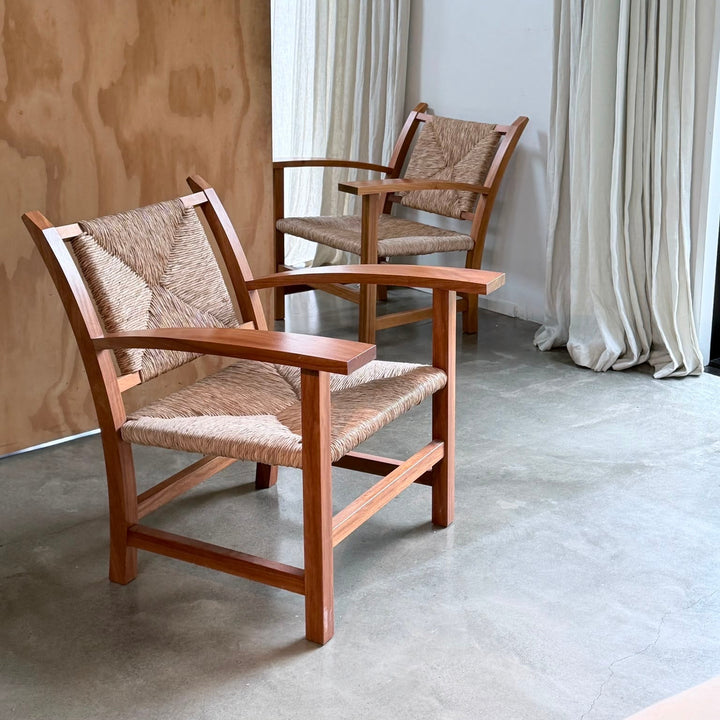 Two mid-century modern wooden armchairs with woven rope seats and backs, positioned in a minimalist room with a plywood wall panel, concrete floor, and cream curtains.
