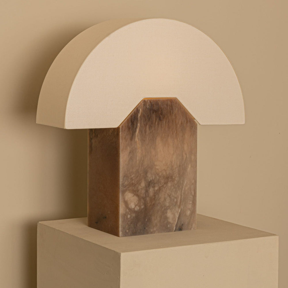 Tobacco table lamp from Shop Dessein Parke featuring a beige semi-circle shade and marble-patterned base, displayed on a beige platform.