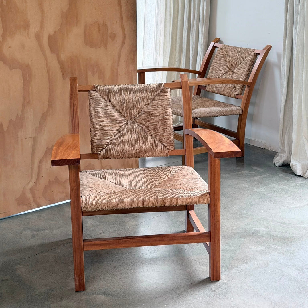 Two mid-century modern wooden armchairs with woven cane seats and backrests, positioned in a room with a plywood panel wall, concrete floor, and off-white curtains.