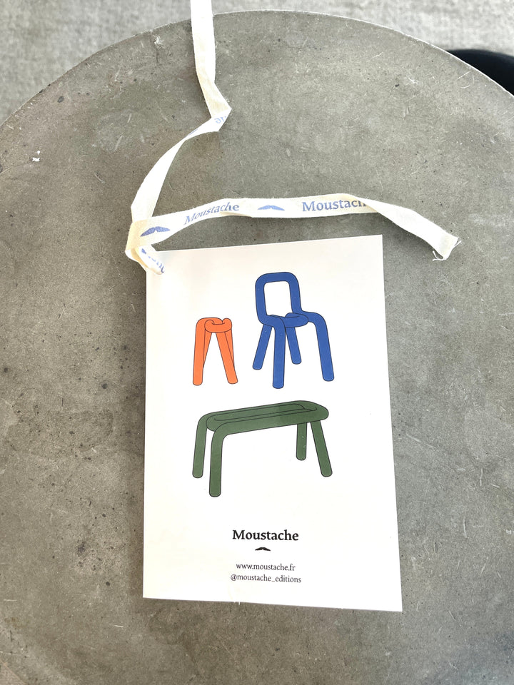 Gray Moustache Bold Chair from Moustache, shown with orange, blue, and green chair illustrations on a white tag with a branded lanyard.
