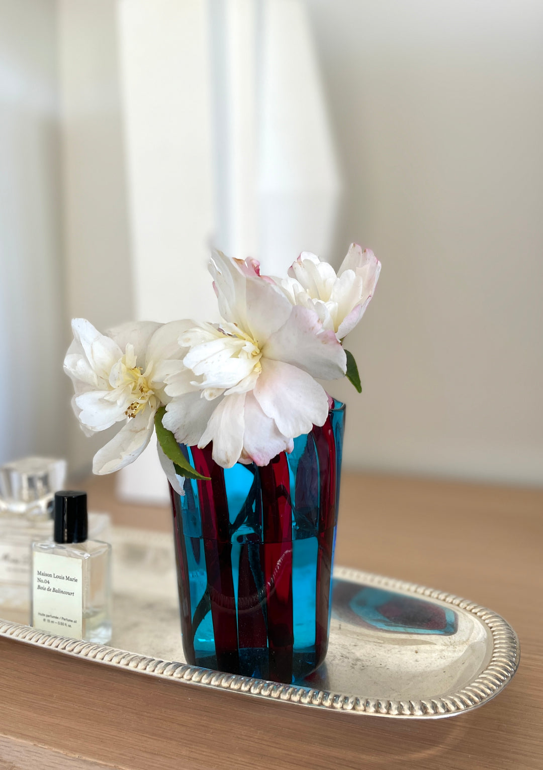 Striped drinking glass by Shop Dessein Parke, filled with white flowers and displayed on a silver tray next to a perfume bottle.