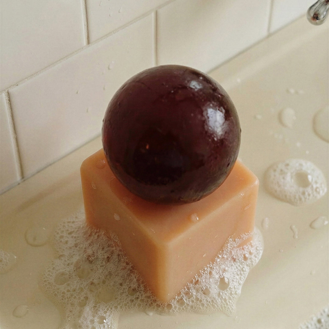 Resin cube  with a dark brown sphere of soap on top, placed on a tiled surface with soap suds.