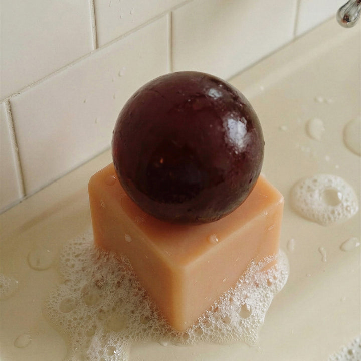 Resin cube  with a dark brown sphere of soap on top, placed on a tiled surface with soap suds.