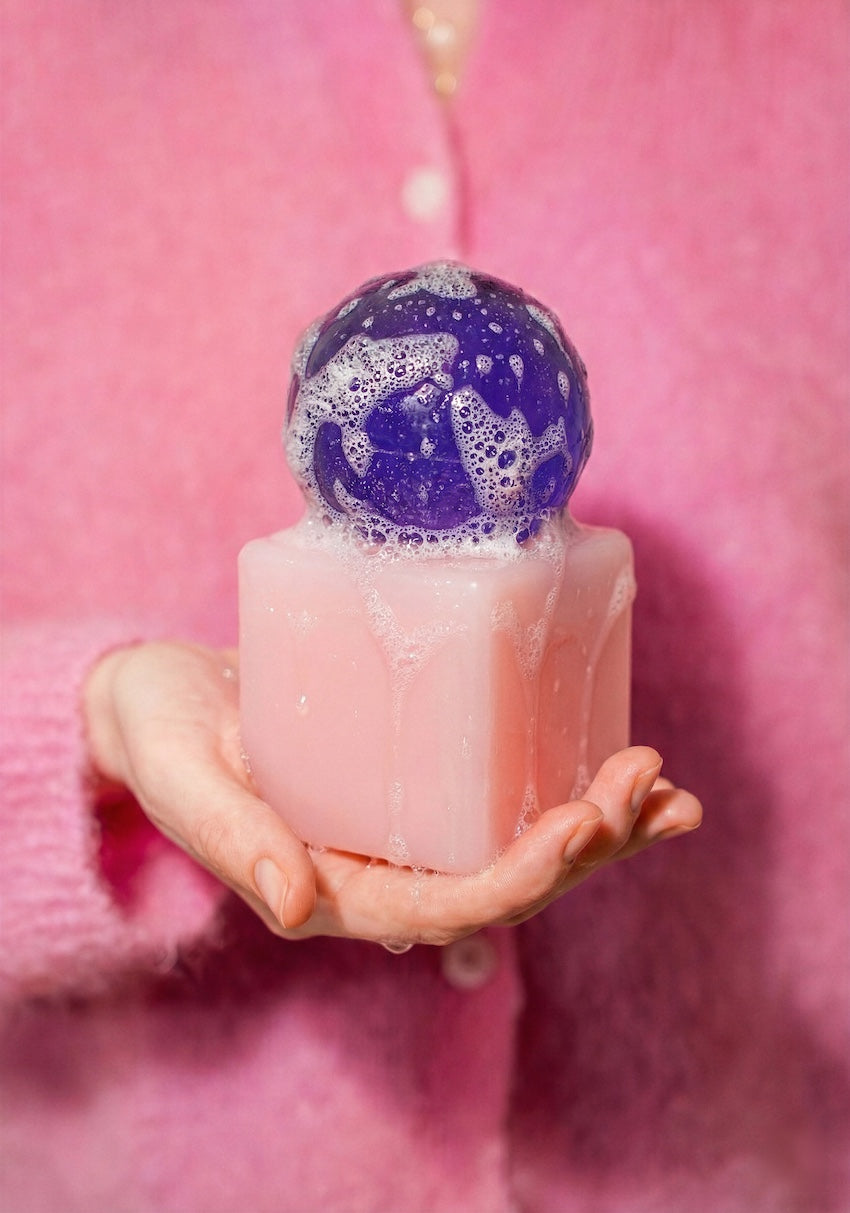 Pink soap bar with a purple bath bomb held in a hand against a pink background