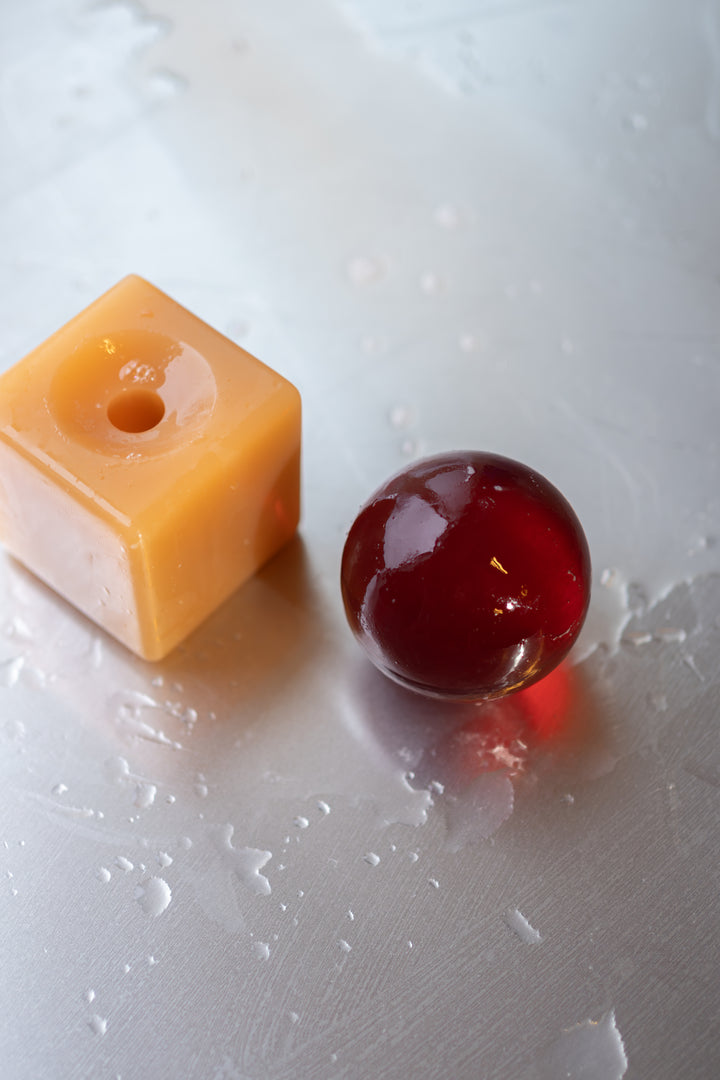 Resin cube  with a dark brown sphere of soap on top, placed on a tiled surface with soap suds.