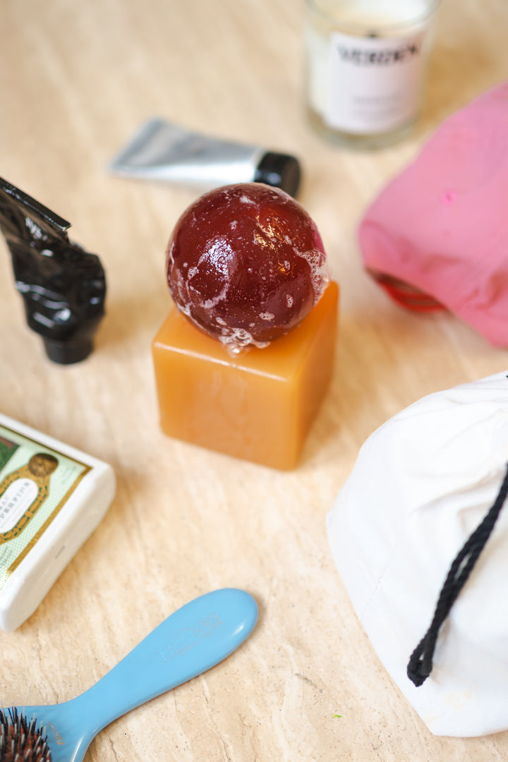 Resin cube with a dark brown sphere of soap on top, on a wooden surface with various hair care items.