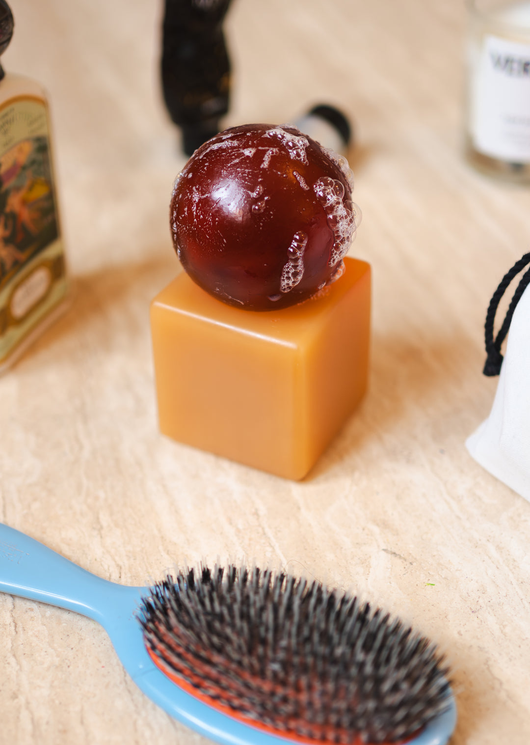 Resin cube with a dark brown sphere of soap on top, on a wooden surface with various hair care items.