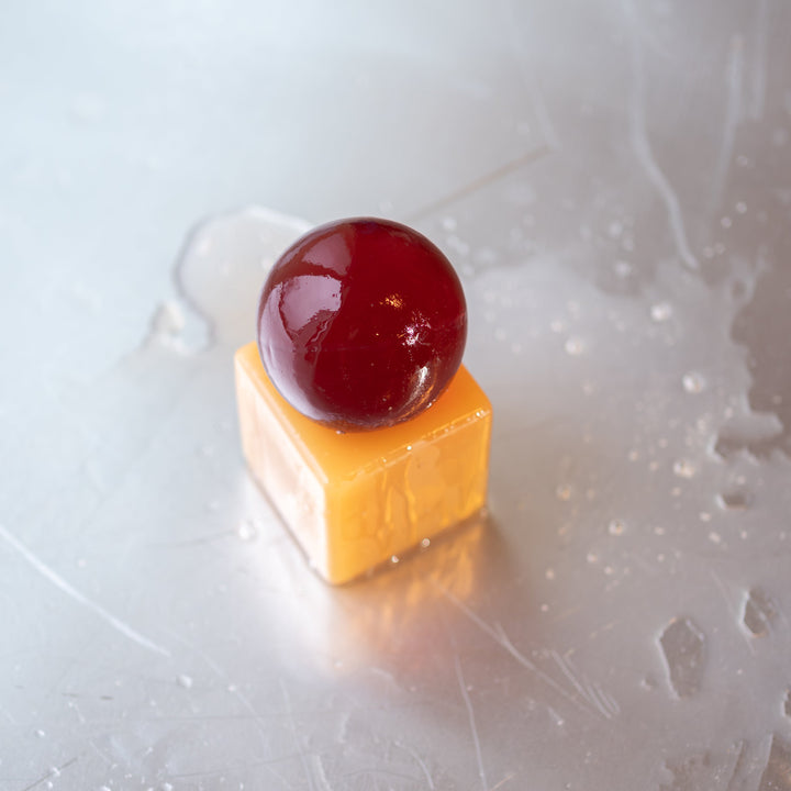 Resin cube  with a dark brown sphere of soap on top, placed on a tiled surface with soap suds.