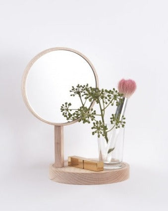 Round wall mirror with a shelf by Moustache Paris, displaying green foliage, a makeup brush, and a lipstick.