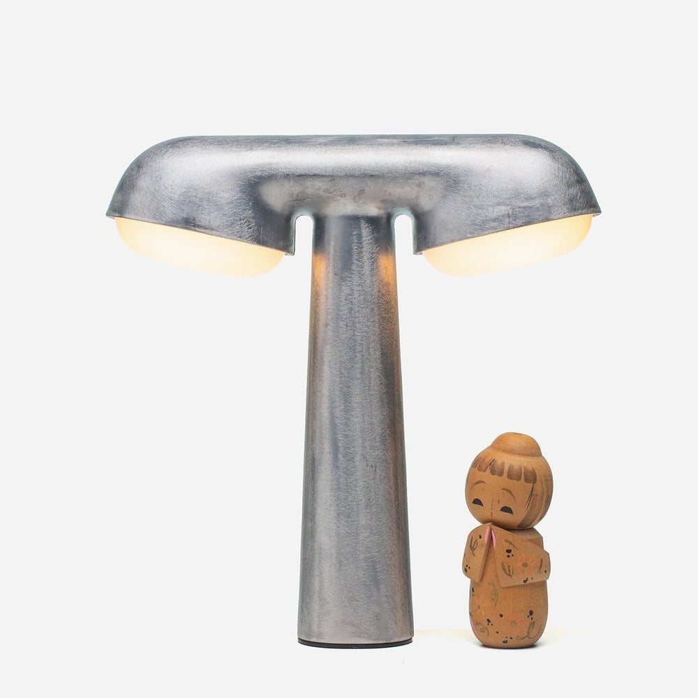 Raw metallic table lamp by Moustache Paris alongside a small, wooden doll, over a white background.
